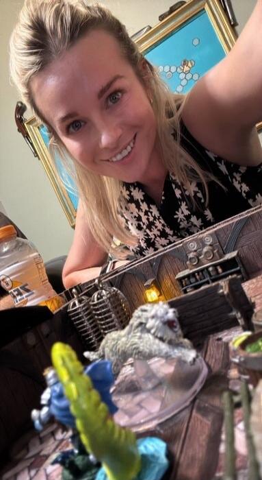 A woman smiling at the camera, with a tabletop role-playing game setup in the foreground.