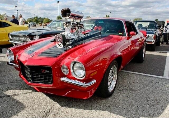 A red classic muscle car with a large blower protruding from the hood, parked at a car show with other vintage cars in the background.