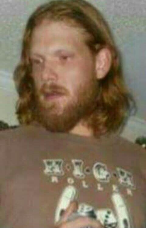 A man with long, wavy brown hair and a beard is looking down. He is wearing a brown t-shirt with some indistinct white graphics and text on the chest.
