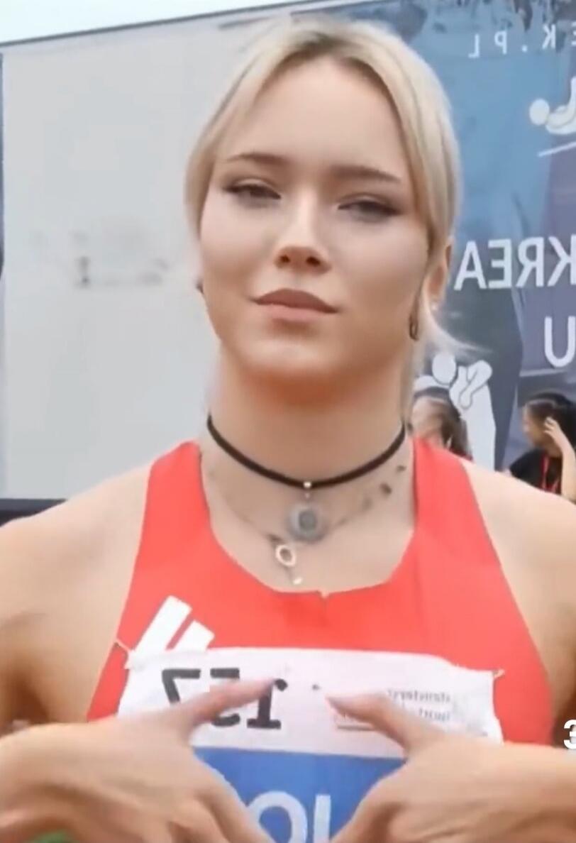 A blonde woman in a red sports top with a bib. Text on the bib includes 'ГГ' and 'OL'. Mirrored Cyrillic text in the background shows 'Я.Б.Г' and 'АЗЯХ U'.
