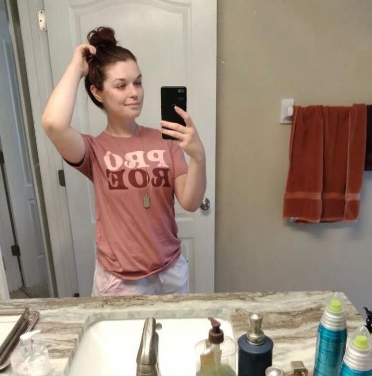 A woman taking a mirror selfie in a bathroom. She is wearing a t-shirt with the text 'PRO BOE' (mirrored as 'BOE BRO') and has her hair in a bun, touching it with one hand. Various bathroom items are visible on the counter.