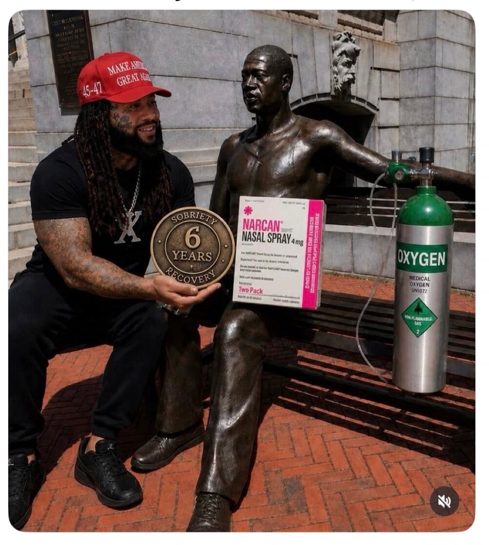 A man wearing a 'MAKE AMERICA GREAT AGAIN 45-47' hat is smiling and holding a medallion that reads 'SOBRIETY 6 YEARS RECOVERY'. He is sitting next to a bronze statue of a man on a bench. The statue is holding a box of 'NARCAN NASAL SPRAY 4mg Two Pack'. An oxygen tank labeled 'OXYGEN MEDICAL OXYGEN UN1072 NON FLAMMABLE GAS 2' is next to the statue.