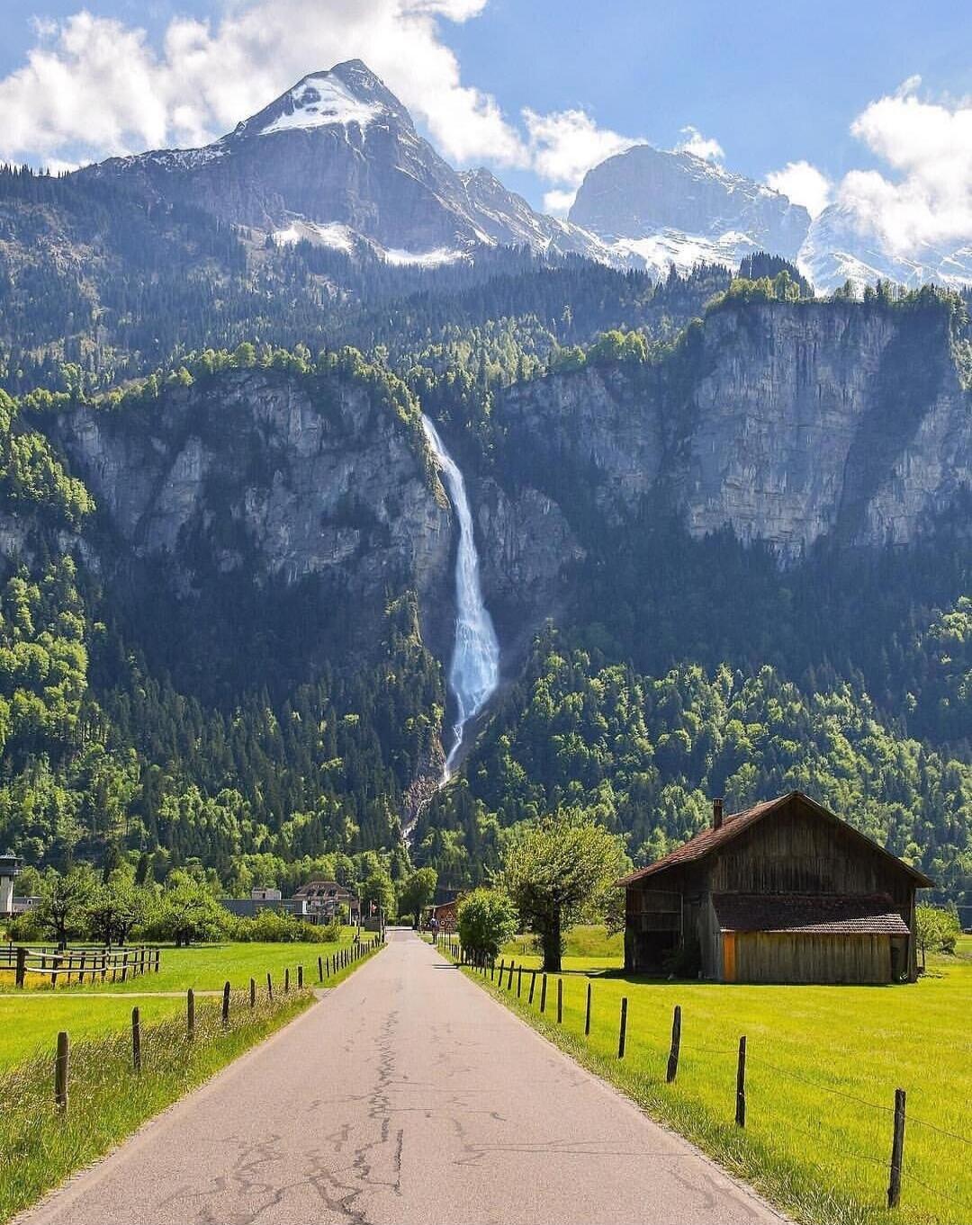 A rural road through a green valley with a tall waterfall cascading down a forested mountain, farm buildings on the right, and snow-capped peaks in the distance.