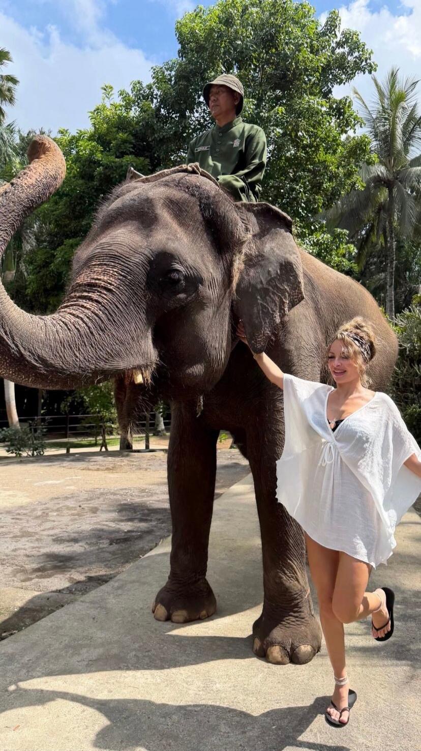A woman in a white cover-up and black bikini top stands next to an elephant, gently touching its ear. An elephant handler is seated on the elephant's back. The scene is outdoors with green trees and a bright sky.