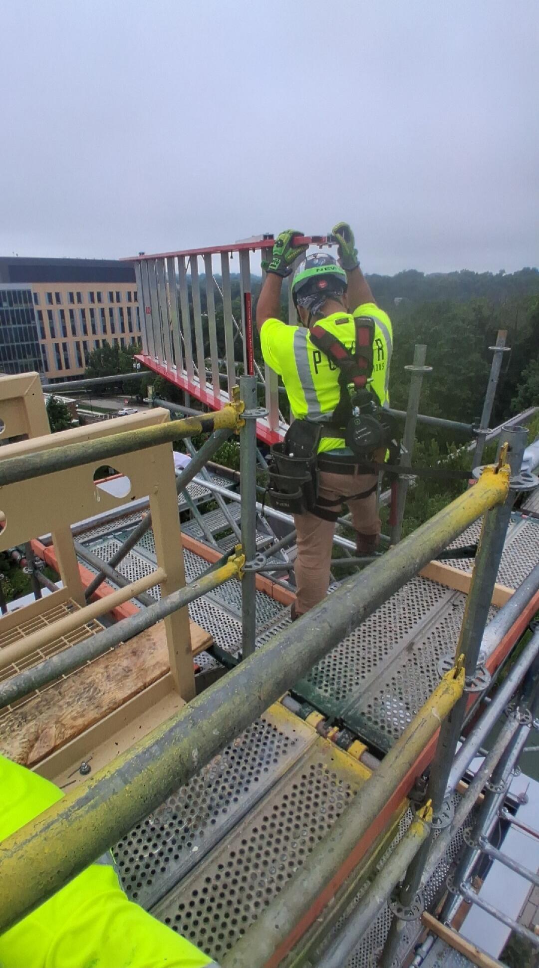 A construction worker in a high-visibility vest and safety harness is on scaffolding, handling a ladder-like structure. Text visible on the helmet says 'NEW', and on the vest 'P R'.