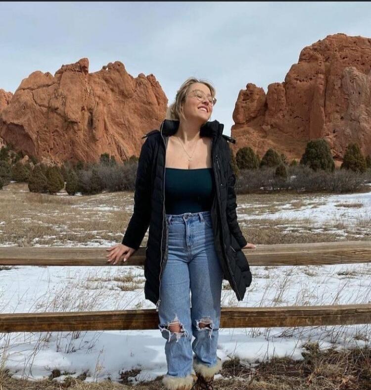 A woman standing outdoors in front of a wooden fence, wearing a black coat, a dark teal top, and ripped jeans with snow on the ground and red rock formations in the background.