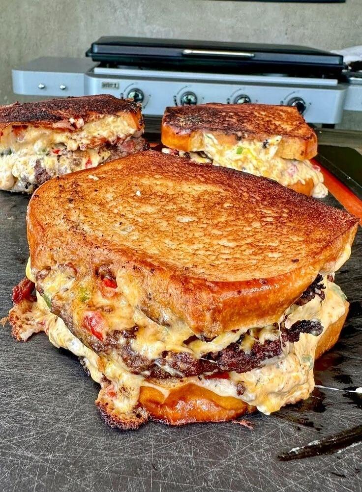 A large, multi-layer grilled cheese sandwich with melted cheese and beef between thick slices of toasted bread. The sandwich appears to be loaded with cheese and other fillings, stacked high on a griddle.