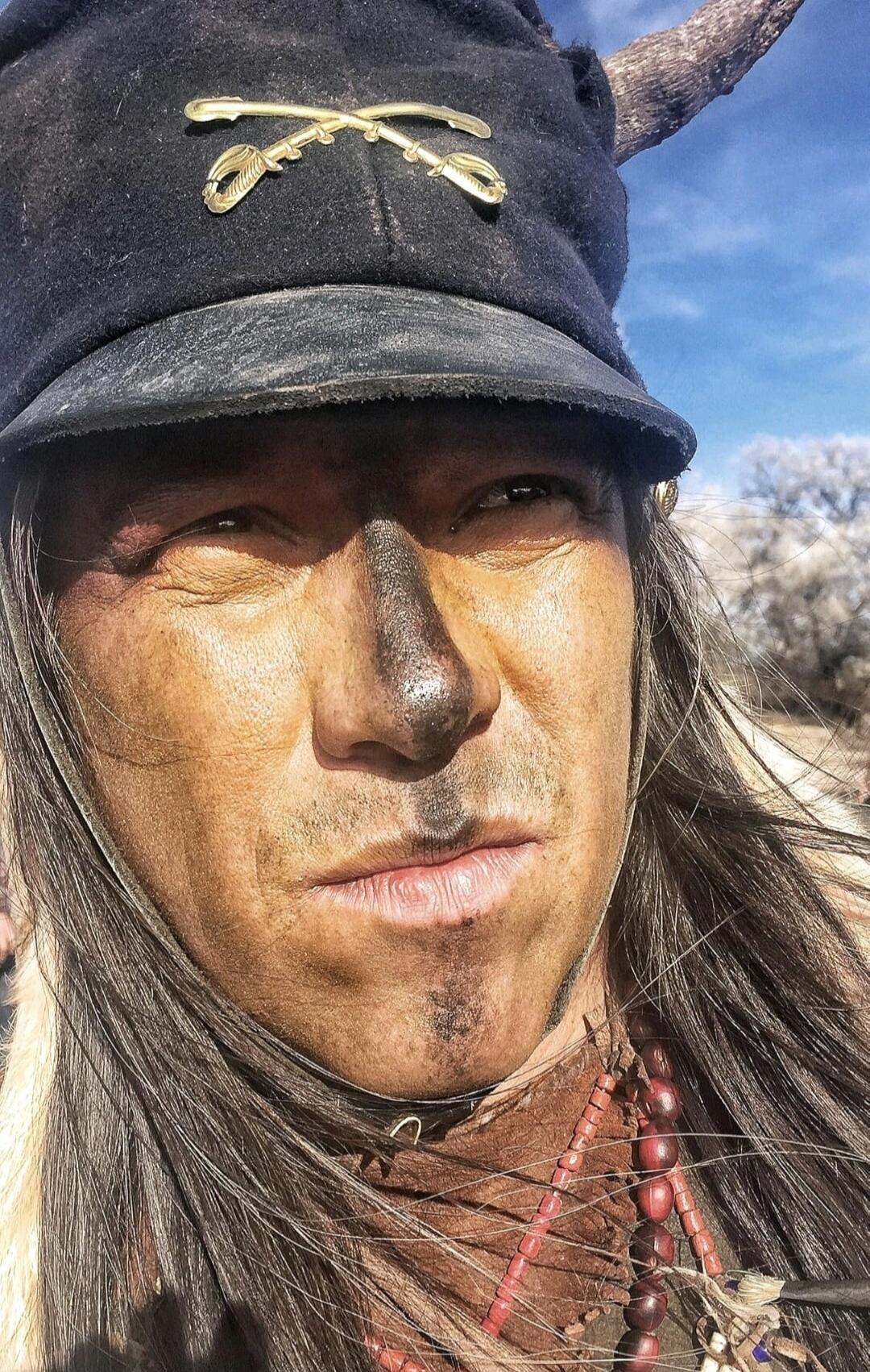 A man with a dark painted face or dirt covering most of his facial skin. He is wearing a dark hat with a decorative emblem and some bead necklaces. Outdoor setting with a blue sky.