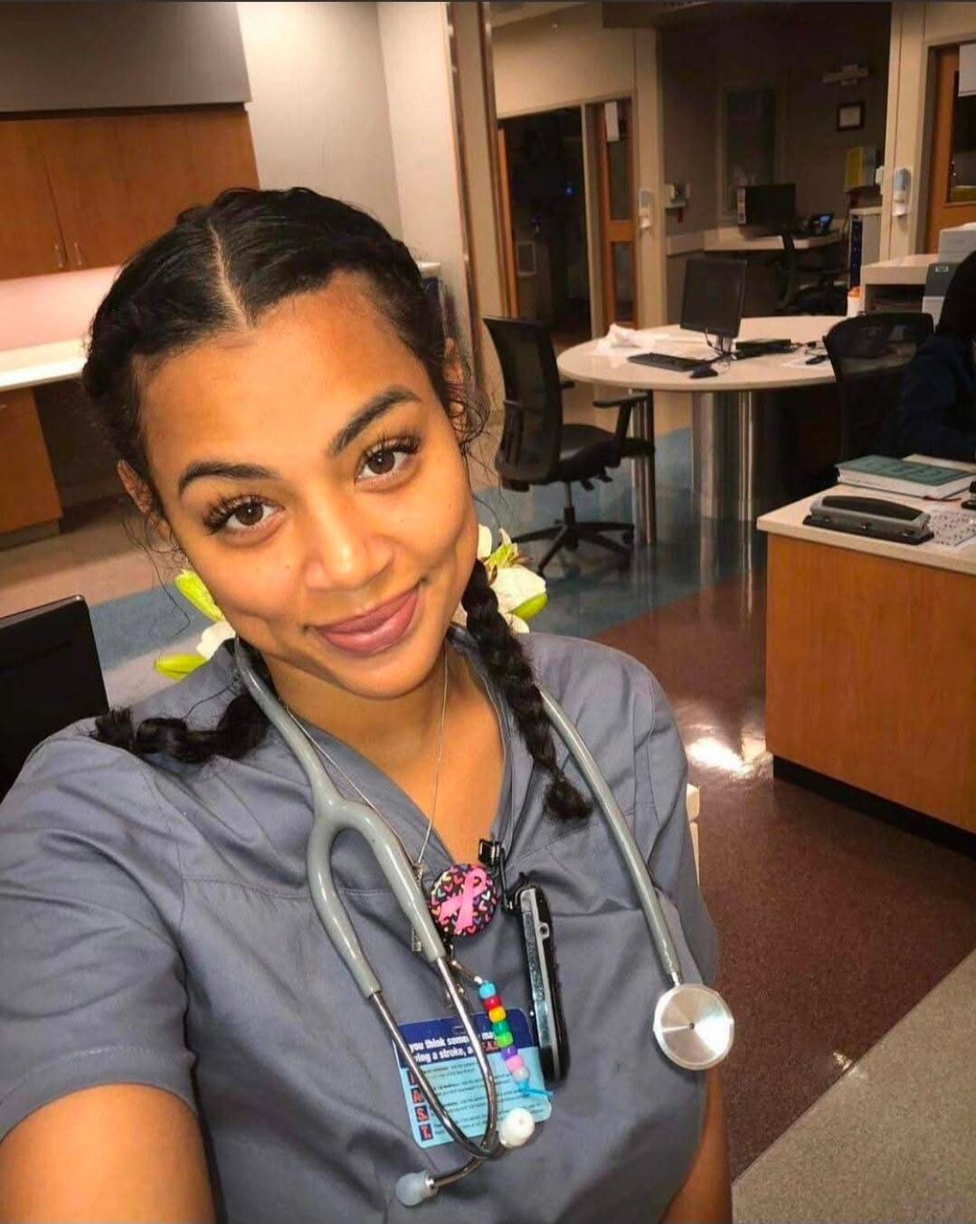 A young female medical professional, possibly a nurse, wearing grey scrubs and a stethoscope, smiling at the camera in what appears to be a hospital or clinic setting. Text on her badge is partially visible, possibly related to medical advice like 'you think some... g a stroke, a...'