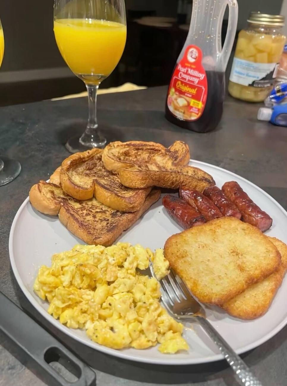 Plate with scrambled eggs, French toast, sausage links, and two slices of buttered toast. A glass of orange juice sits behind the plate. In the background, a bottle of syrup labeled Original and a jar of pickles are visible.