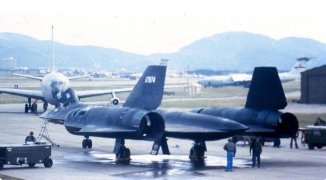 Two Lockheed SR-71 Blackbird reconnaissance aircraft are parked on an airfield. One aircraft has the tail number 0974 visible on its vertical stabilizer. Other planes and mountains are in the background.