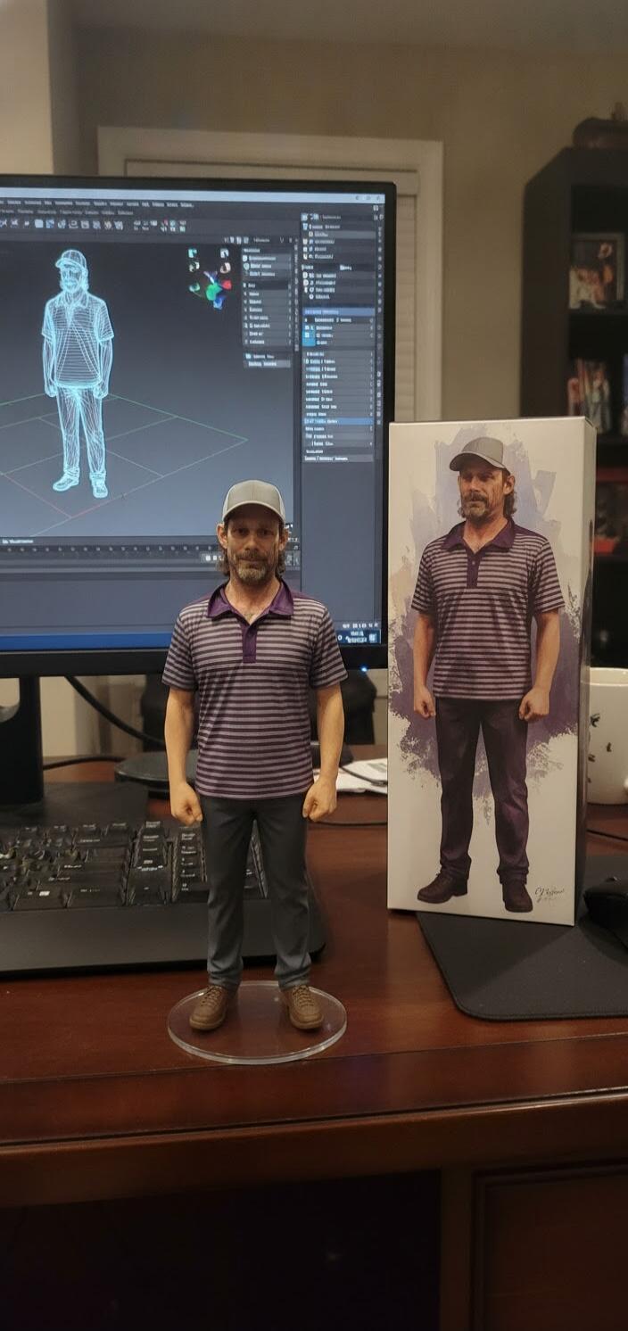 A 3D printed figurine of a man stands on a wooden desk, wearing a striped polo shirt, dark pants, and a baseball cap. Behind it, a computer monitor displays a wireframe 3D model of the same man, indicating the digital design process. To the right, a box with an artistic illustration of the man is visible, suggesting packaging for the figurine.