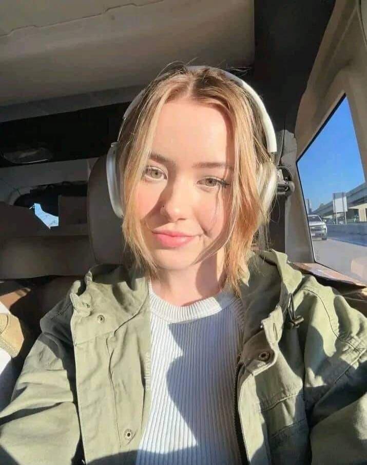 A young woman with blonde hair and headphones is smiling at the camera from inside a car. Sunlight illuminates her face. There's a faint watermark '@aluraye' on her jacket.