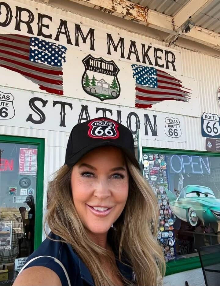 DREAM MAKER STATION HOT ROD HIGHWAY TEXAS ROUTE 66 ROUTE 66 OPEN. A woman is smiling at the camera, wearing a black cap with a Route 66 logo. She is standing in front of a building with various signs related to Route 66 and hot rods.