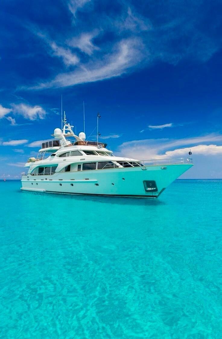 A luxurious white yacht floats on crystal clear turquoise water under a bright blue sky with scattered clouds.