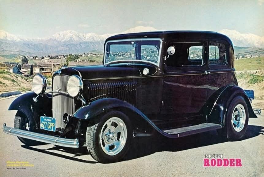 A vintage black hot rod car, possibly a Ford, is parked on a road with mountains and a town in the background. The license plate reads 'CALIFORNIA 627 GPP'. The bottom right corner has text 'STREET RODDER'.