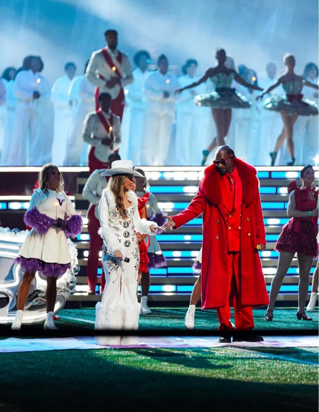 A live stage performance with a diverse group of dancers and singers in the background. A man in a bright red coat shakes hands with a woman wearing a white dress and a large white hat, as other performers in white and colorful outfits stand and dance on a lit, multi-tiered stage.