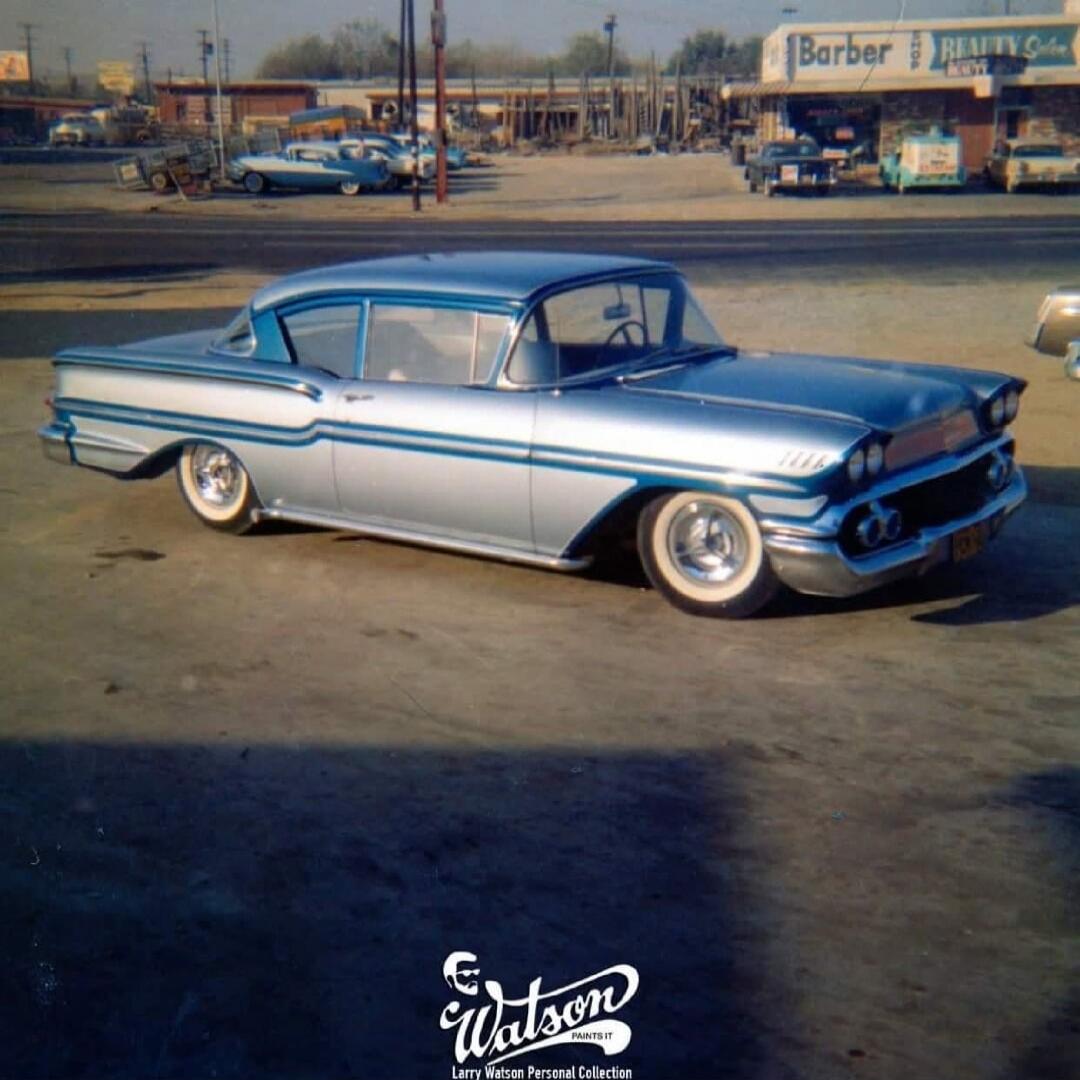 A vintage silver car with blue pinstriping, lowered suspension, and white-wall tires is parked on asphalt. In the background, there are other cars and a building with a sign that reads 'Barber Realty Sales'. The bottom of the image features a 'Watson PAINTS IT' logo and 'Larry Watson Personal Collection'.