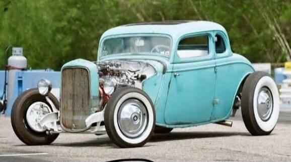 A vintage light blue hot rod car with an exposed engine and white wall tires, parked outdoors.
