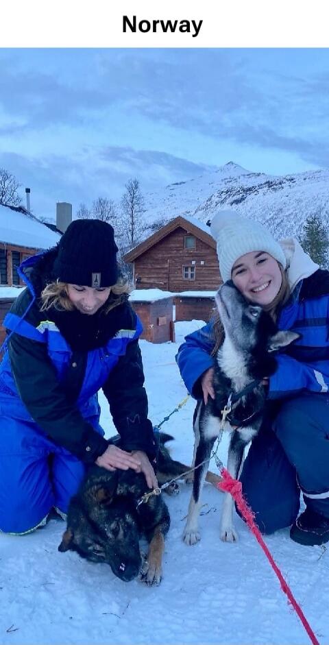 Norway. Two women are petting dogs in a snowy landscape with mountains and wooden cabins in the background. They are dressed in winter gear.