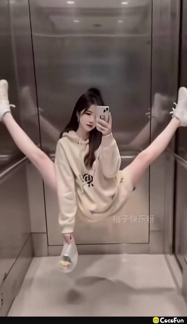 A person performing a wide middle split in an elevator while taking a selfie, wearing a beige hoodie and white socks.