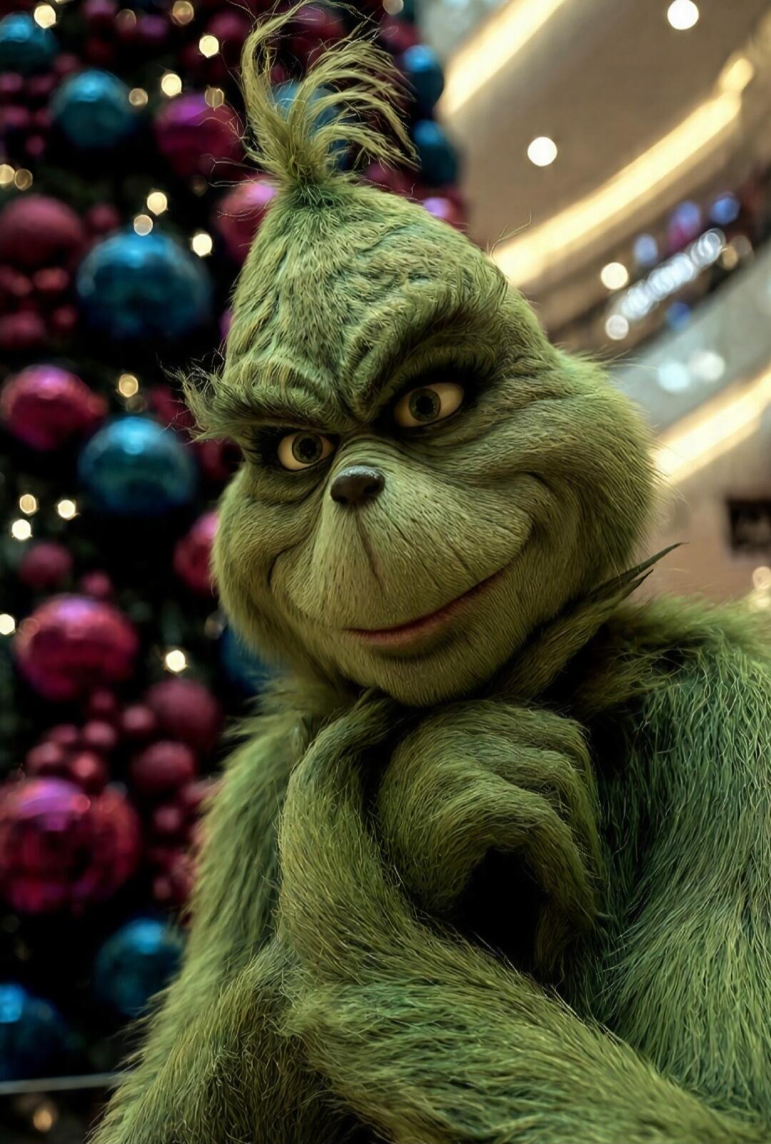 A green Grinch-like character posing with hands together in front, festive decorations in the background.