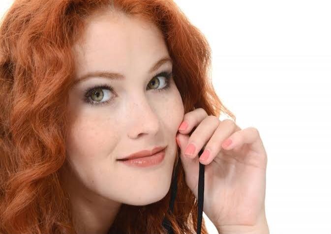 A close-up shot of a young woman with vibrant red curly hair and green eyes. She has a light complexion with freckles across her nose and cheeks. Her lips are slightly parted, and she is looking towards the camera with a gentle smile. Her right hand is raised, with her fingers delicately touching her face near her ear, holding a thin black strap. T