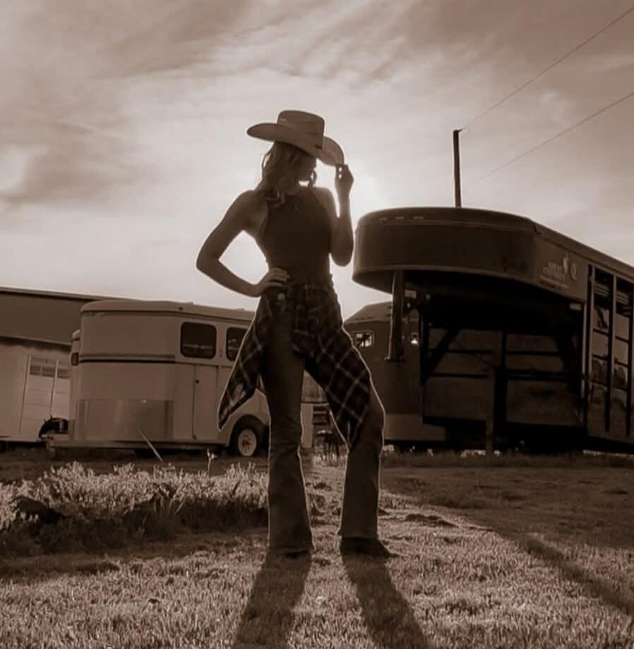 A woman in a cowboy hat, tank top, and jeans with a plaid shirt tied around her waist, posing outdoors at sunset with horse trailers in the background. The image has a sepia tone.