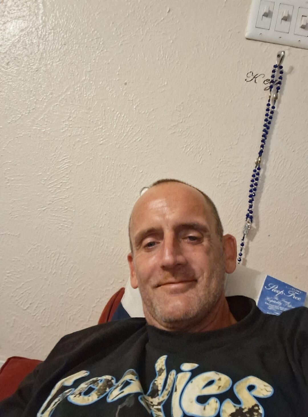 A man taking a selfie, smiling slightly. He has short hair and a beard, wearing a dark t-shirt. In the background, there's a textured wall, a light switch, a blue rosary, and a tag that says 'Sleep-Free'.