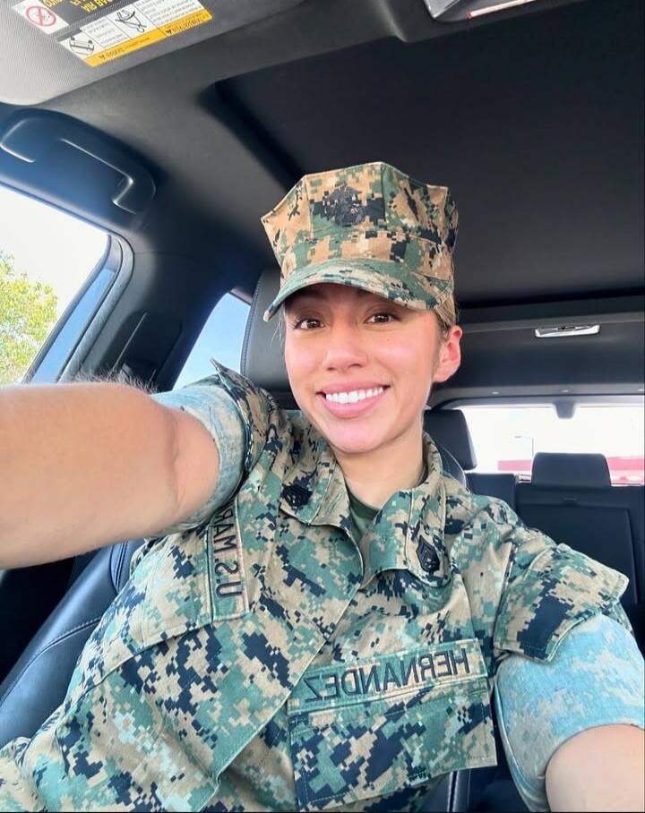 A woman in a US Marine Corps uniform is taking a selfie in a car. She is smiling. Text on uniform includes 'U.S. MARINES' and 'HERNANDEZ'.