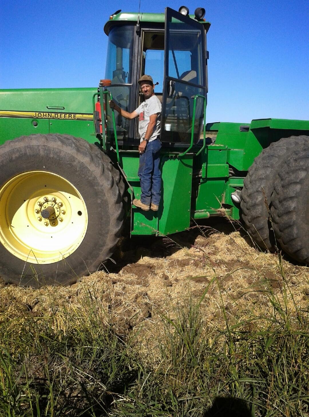 A man in a hat and t-shirt stands on the steps of a large green John Deere tractor in a field under a clear blue sky. The tractor has 'JOHN DEERE' written on its side.