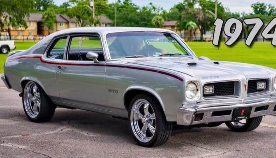 A silver 1974 Pontiac GTO classic car with custom chrome wheels and a red stripe, parked outdoors. Text on image: 1974, GTO.
