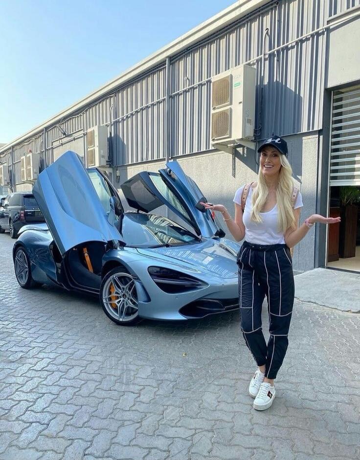 A blonde woman in a white t-shirt, black pants, and a baseball cap stands next to a light blue McLaren 720S with its butterfly doors open. She is smiling and gesturing towards the car.