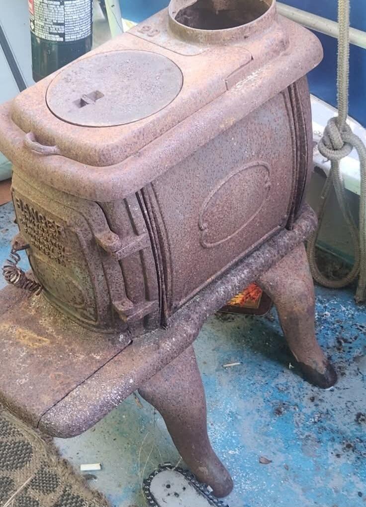 Old rusty cast-iron stove on a blue boat deck, mounted on a metal stand with curved legs. The surface is covered in rust and dust.