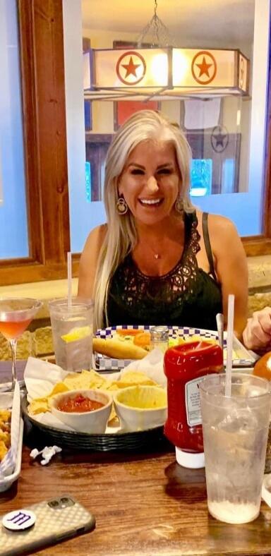 A woman with blonde hair is smiling at a table in a restaurant, with food and drinks in front of her. There are chips, dips, and a hot dog or sandwich on the table. A cocktail and a glass of water are also visible.