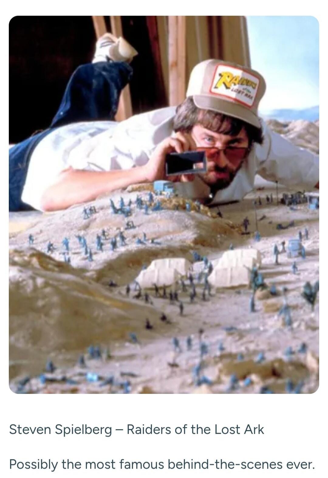 Steven Spielberg - Raiders of the Lost Ark. Possibly the most famous behind-the-scenes ever. A man, Steven Spielberg, wearing a hat with 'RAIDERS OF THE LOST ARK' written on it, is lying down and looking through a small viewfinder at a miniature desert set with many small blue and grey figures, possibly representing soldiers or characters, and some