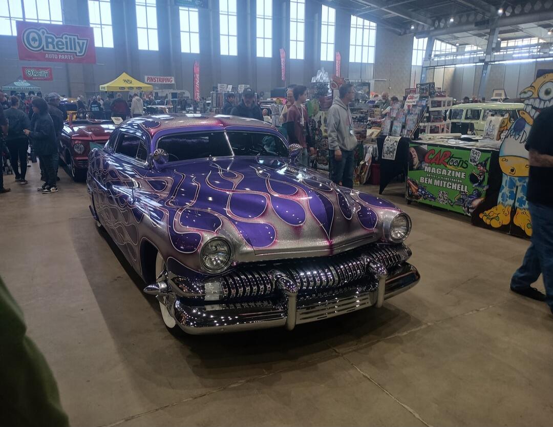A purple vintage car with purple flame decals on display at an indoor car show. There are people walking around and vendor stalls in the background; a banner with 'O'Reilly' is visible.
