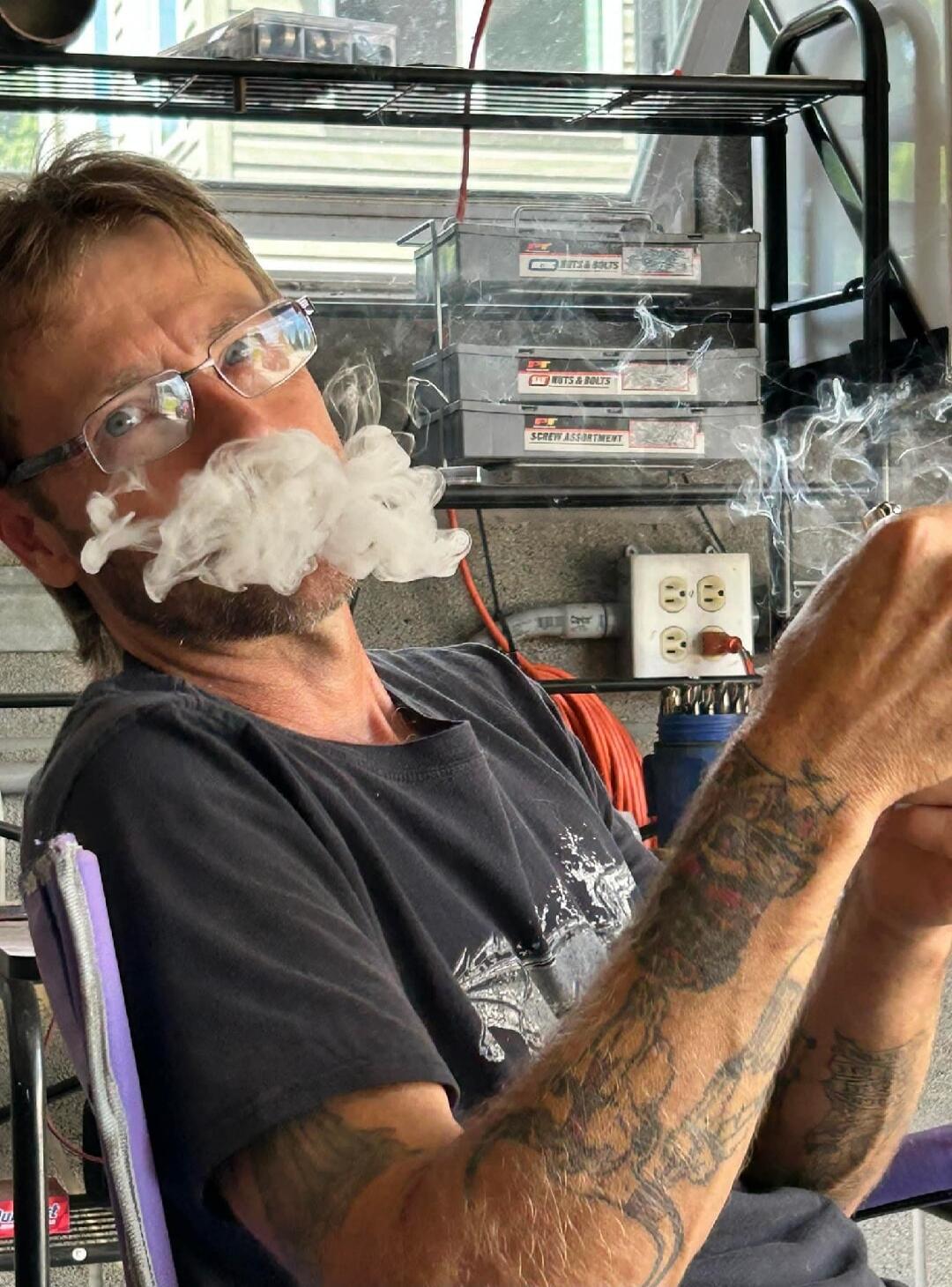 A man with glasses and tattoos exhales a large cloud of smoke, forming a mustache shape, in what appears to be a garage or workshop. Storage bins labeled 'NUTS & BOLTS' and 'SCREW ASSORTMENT' are visible in the background.