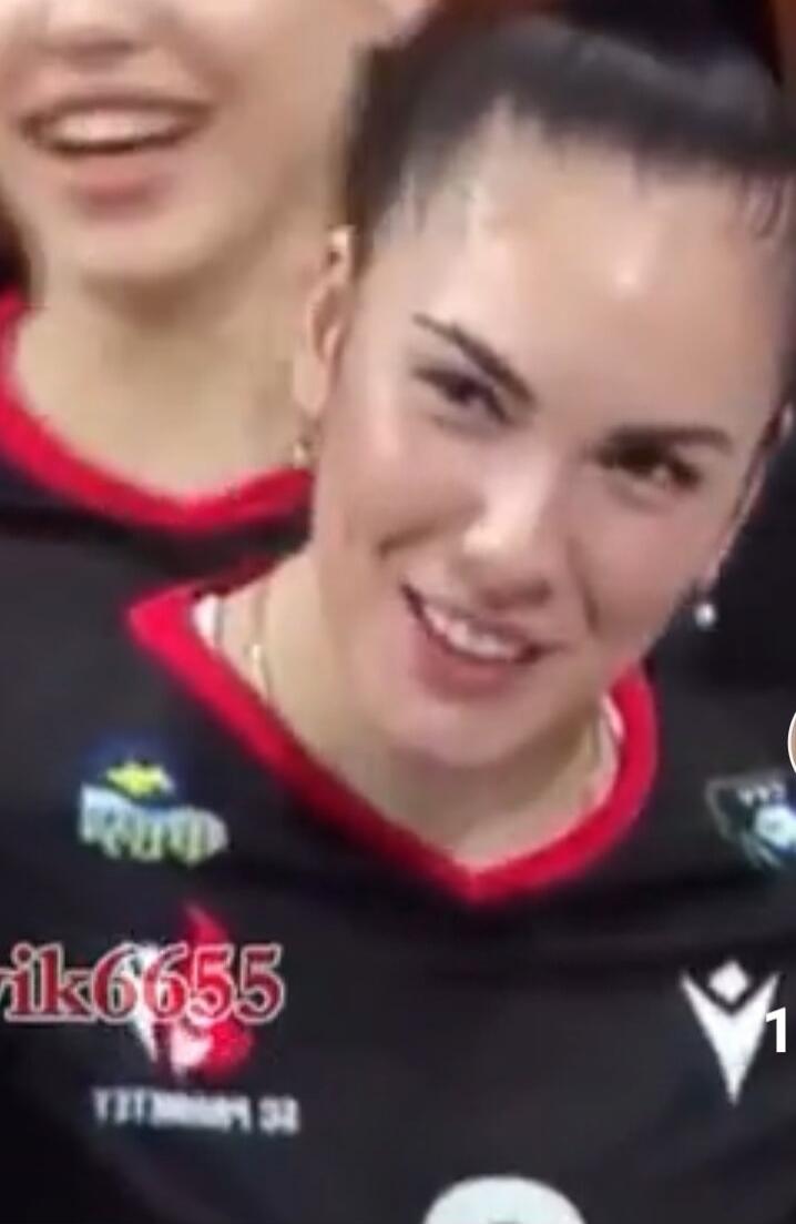 A woman in a sports uniform is smiling or smirking, with another person smiling in the background. There is a watermark 'rik6655' visible on the uniform.