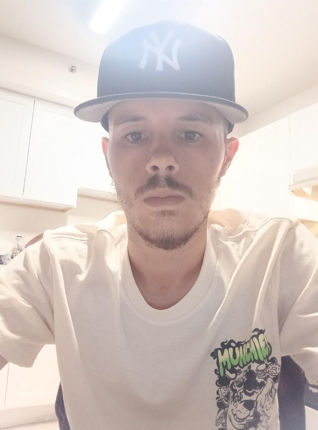A man wearing a black baseball cap with a white 'NY' logo and a white t-shirt with a 'MUNCHIES' graphic print. He has a beard and mustache.