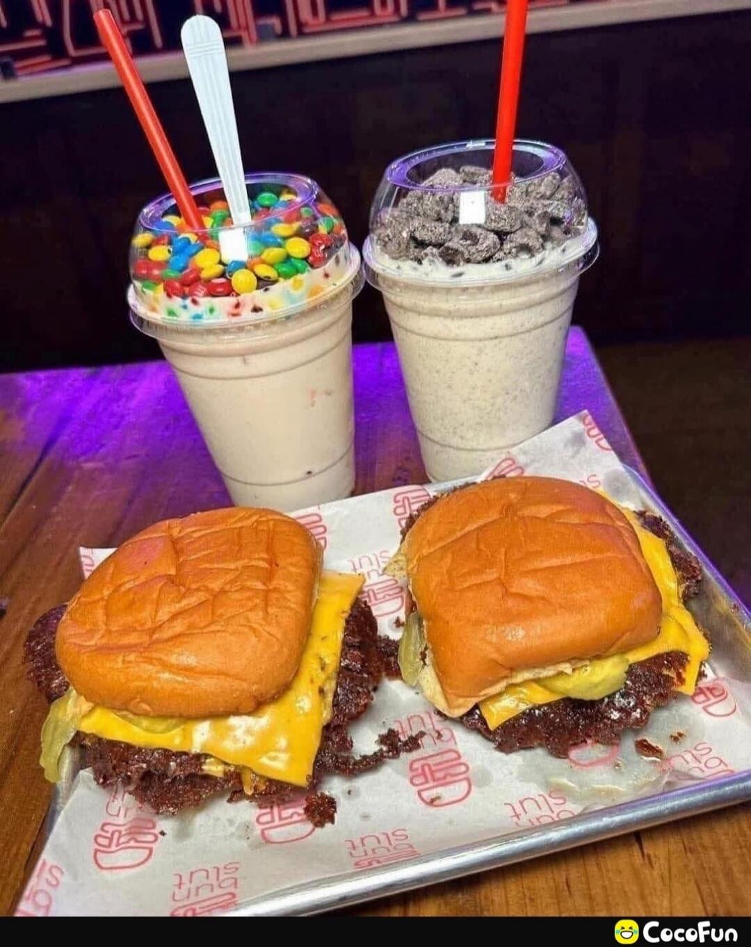 Two smash burgers with melted cheese and pickles on a tray, accompanied by two milkshakes. One milkshake is topped with colorful M&M's, and the other with dark cookie crumbs. The paper lining the tray has the text 'BUN SLUT' printed on it.