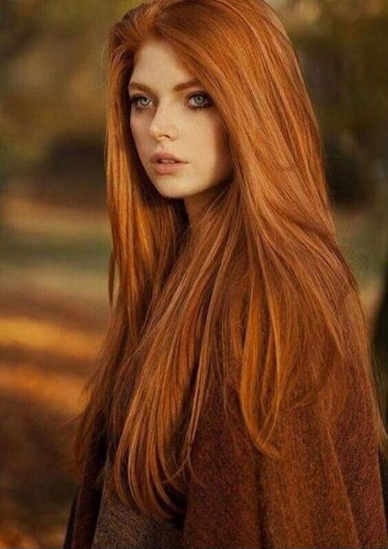 A beautiful young woman with long, flowing red hair and striking blue eyes, looking thoughtfully into the distance. She is wearing a warm, brown cloak, and the background suggests an autumnal setting with blurred trees and warm, golden light.