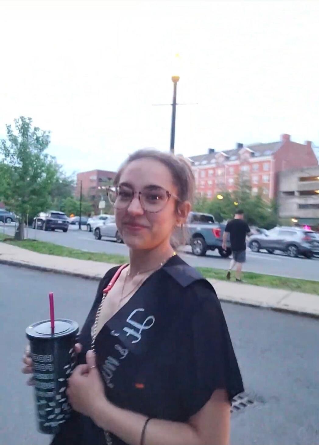 A young woman wearing glasses and a black sash, holding a dark cup with a pink straw, stands on a street. The sash has partially visible text 'f Mo'. Cars and buildings are in the background.