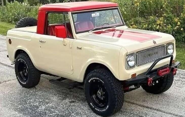 A vintage International Harvester Scout SUV/pickup truck, cream-colored with a red roof and red stripes on the hood, parked outdoors. It features large off-road tires and a front bumper with a winch.