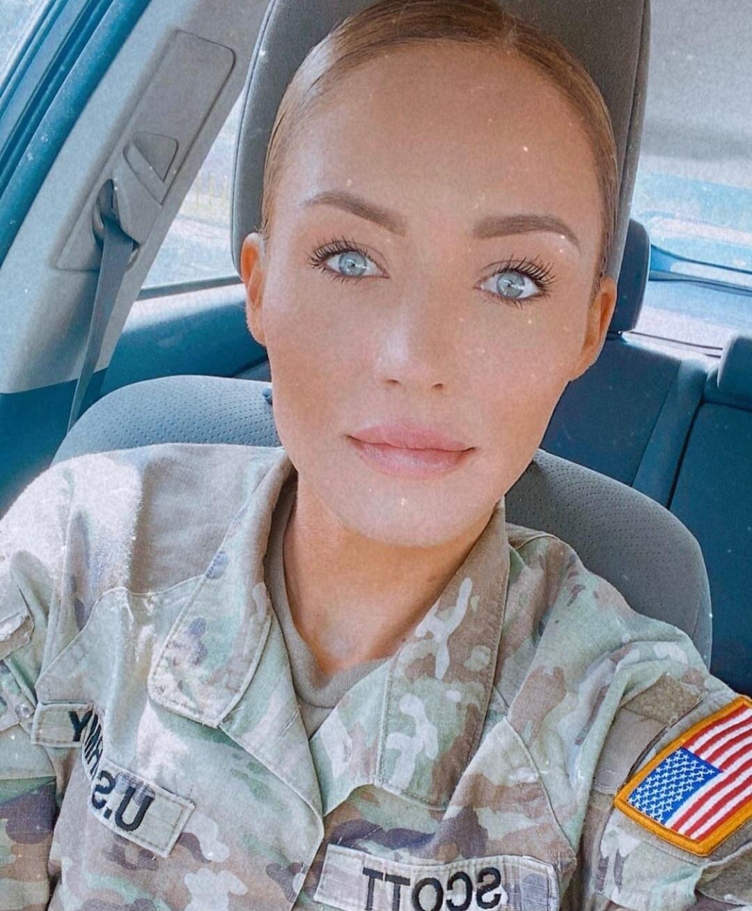 A woman in a US Army uniform, with 'U.S.' and 'SCOTT' visible on her patches, and an American flag on her sleeve, is taking a selfie in a car.
