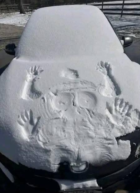 A car covered in snow with an impression of a face and hands on the hood/windshield.