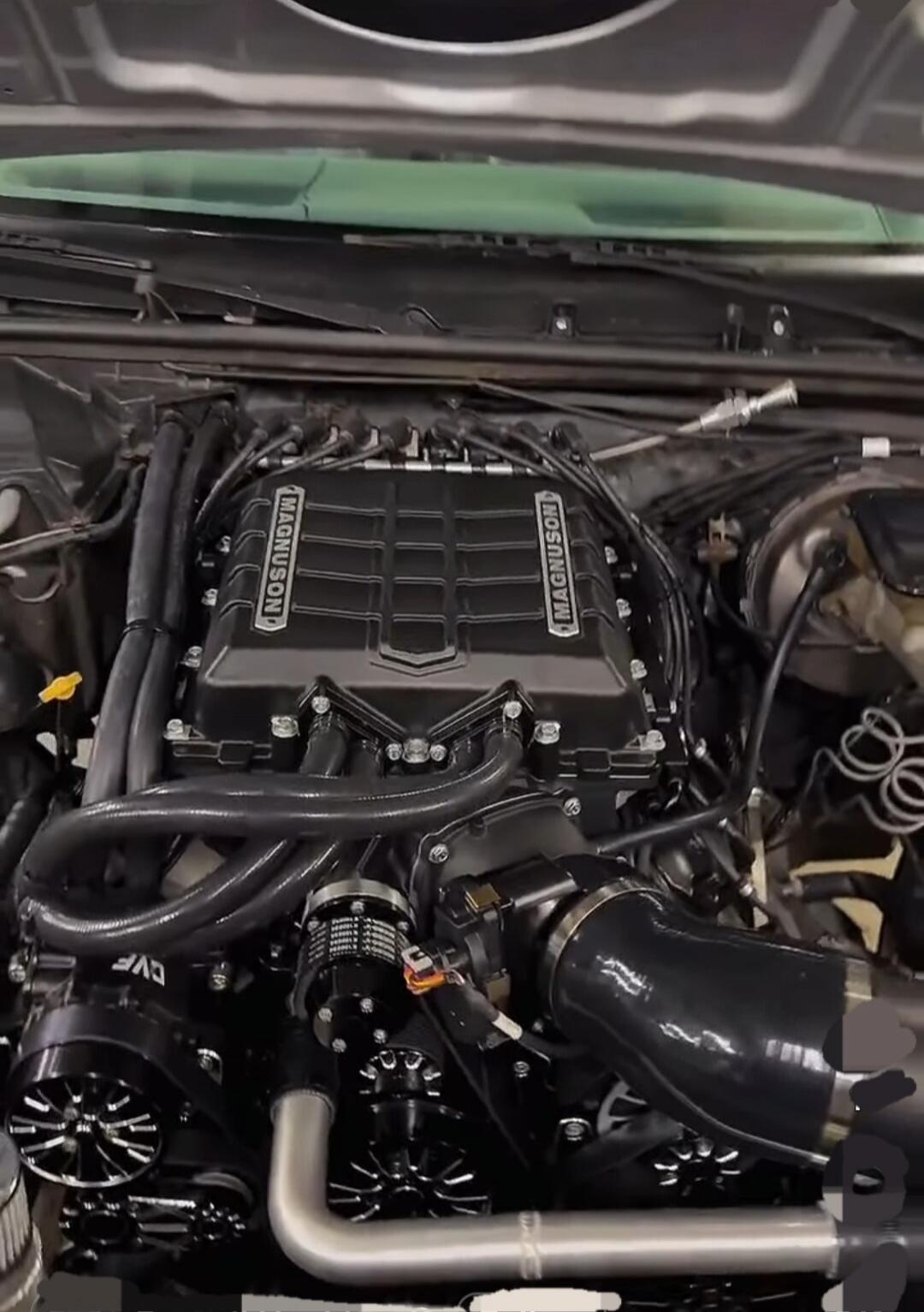 Under-hood view of a car engine with a prominent black intake/plenum and various belts and hoses.