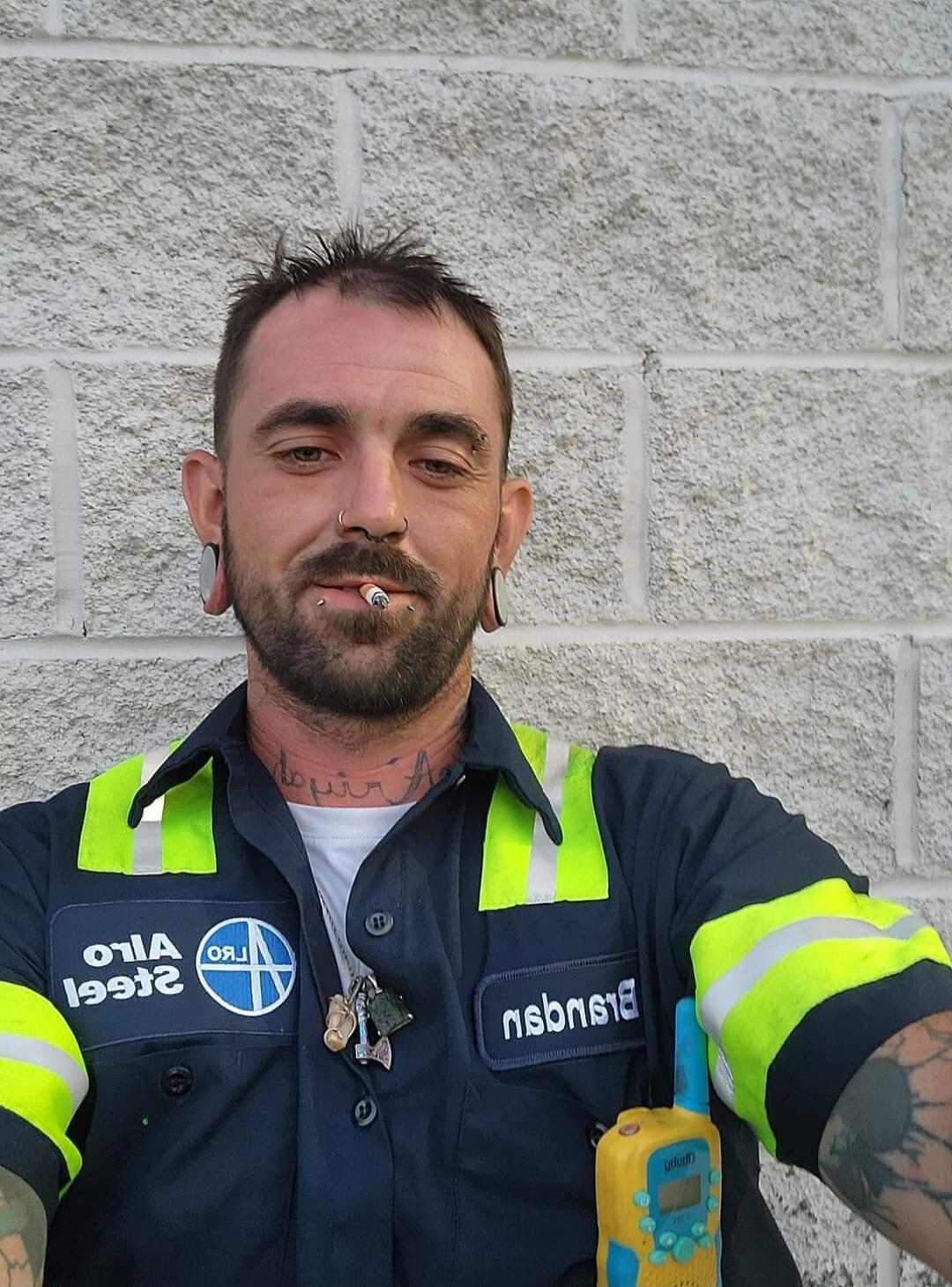 A man with a beard, piercings, and tattoos is smoking a cigarette. He is wearing a work uniform with reflective stripes. Text on uniform: 'Alro Steel', 'Brendan'. He has a walkie-talkie clipped to his shirt.