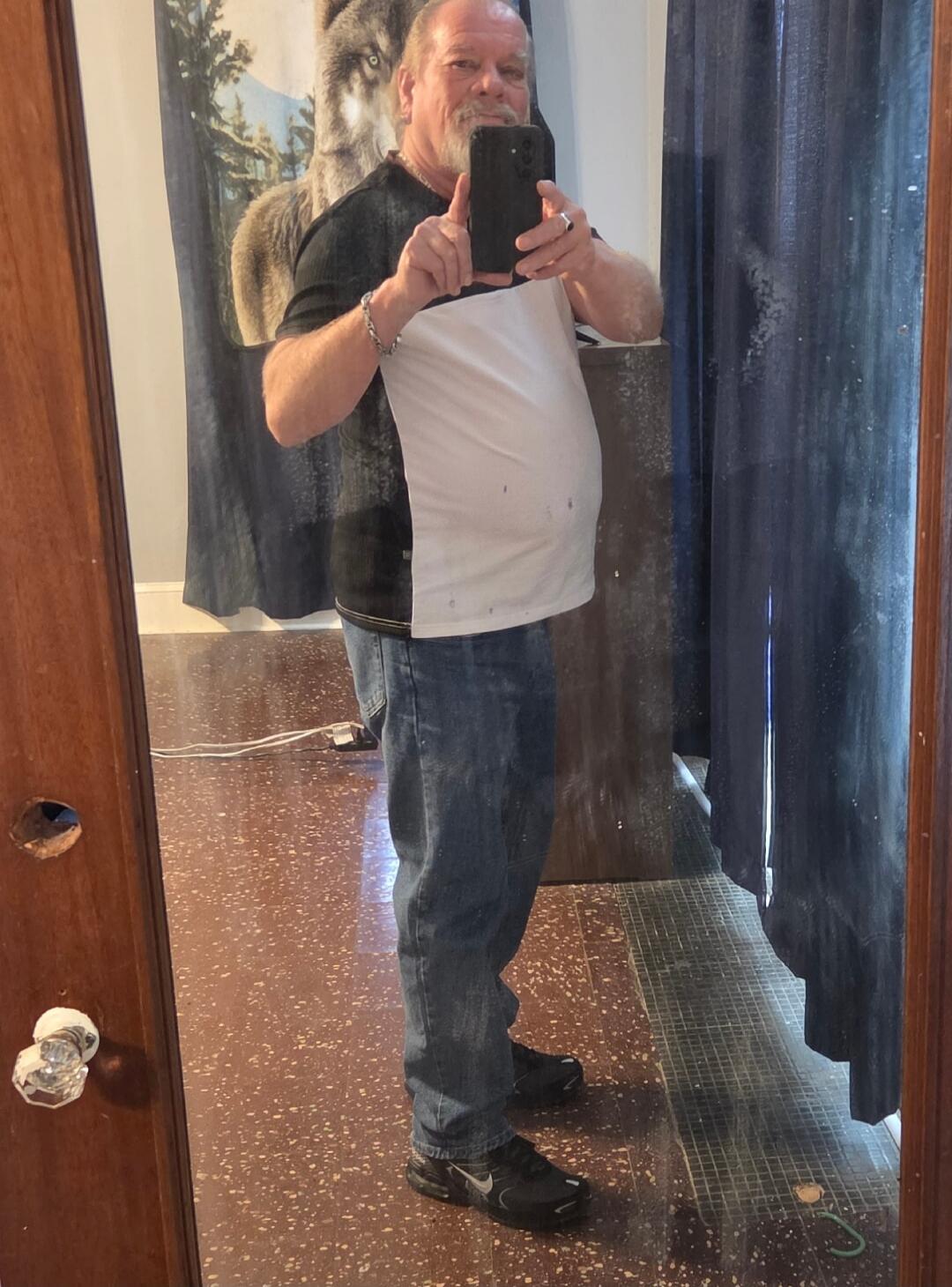 A man with a beard takes a mirror selfie, wearing a black and white t-shirt, blue jeans, and black sneakers. A wolf tapestry is visible in the background.