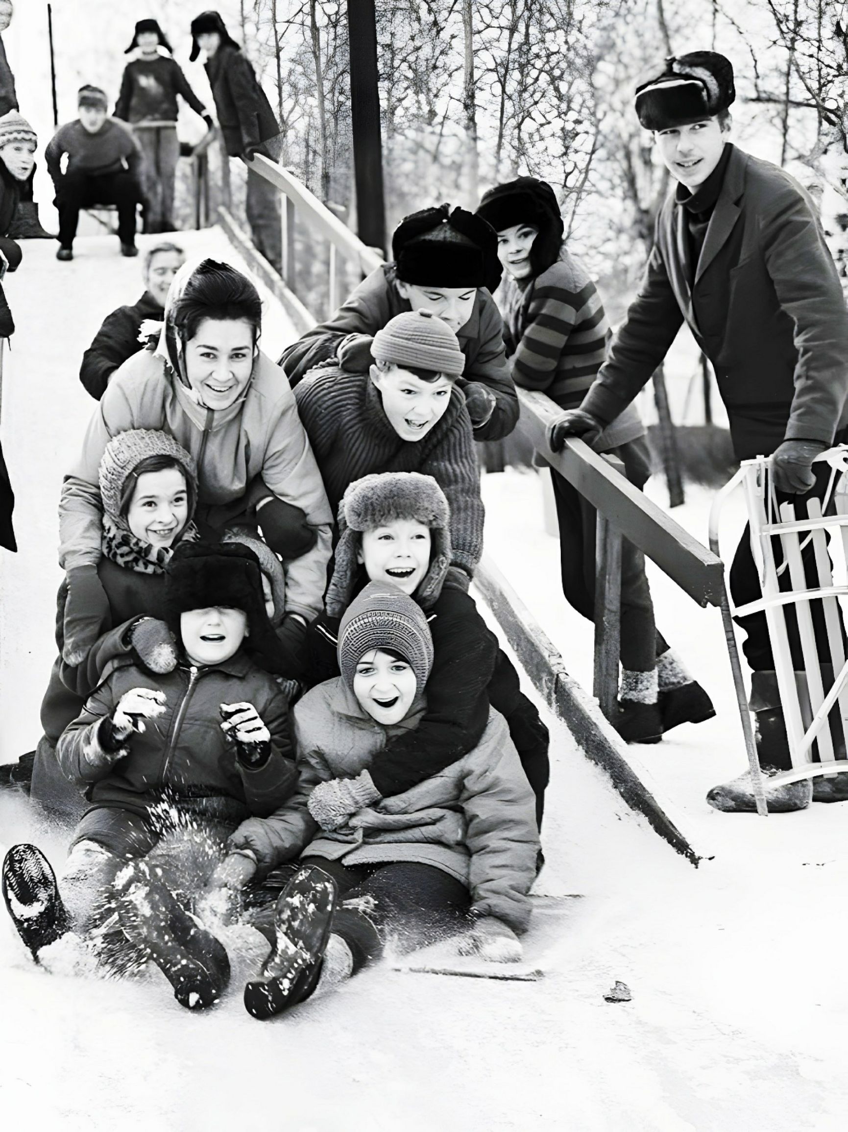 Группа взрослых и детей на санках на заснеженном склоне; все улыбаются.